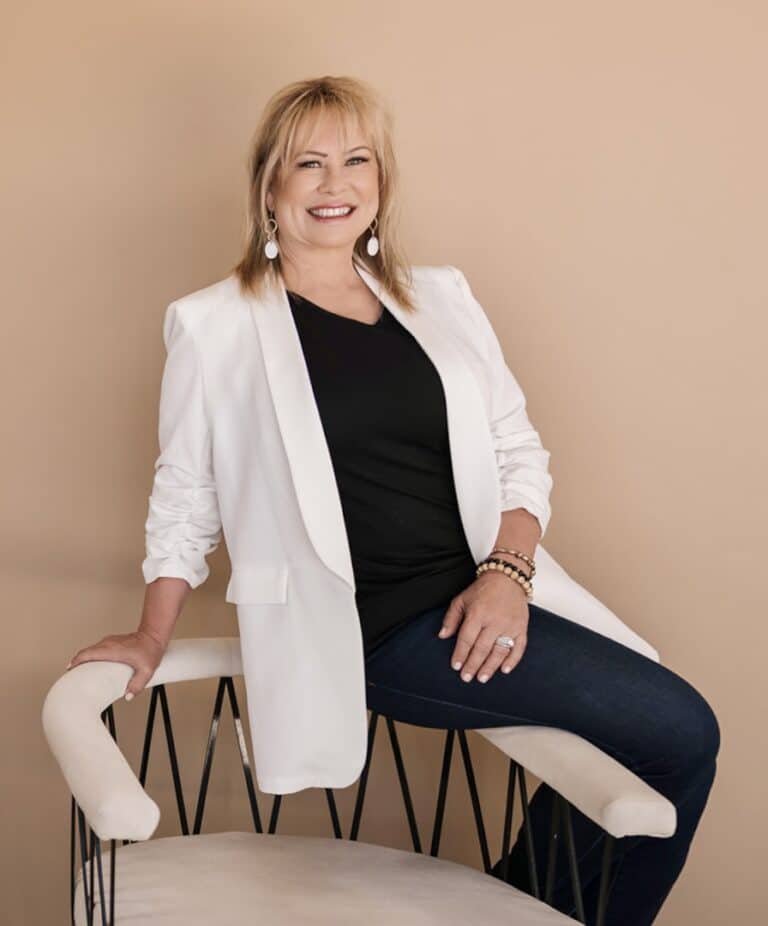 wina gill siting on a white chair with a white jacket and black pants and a black top